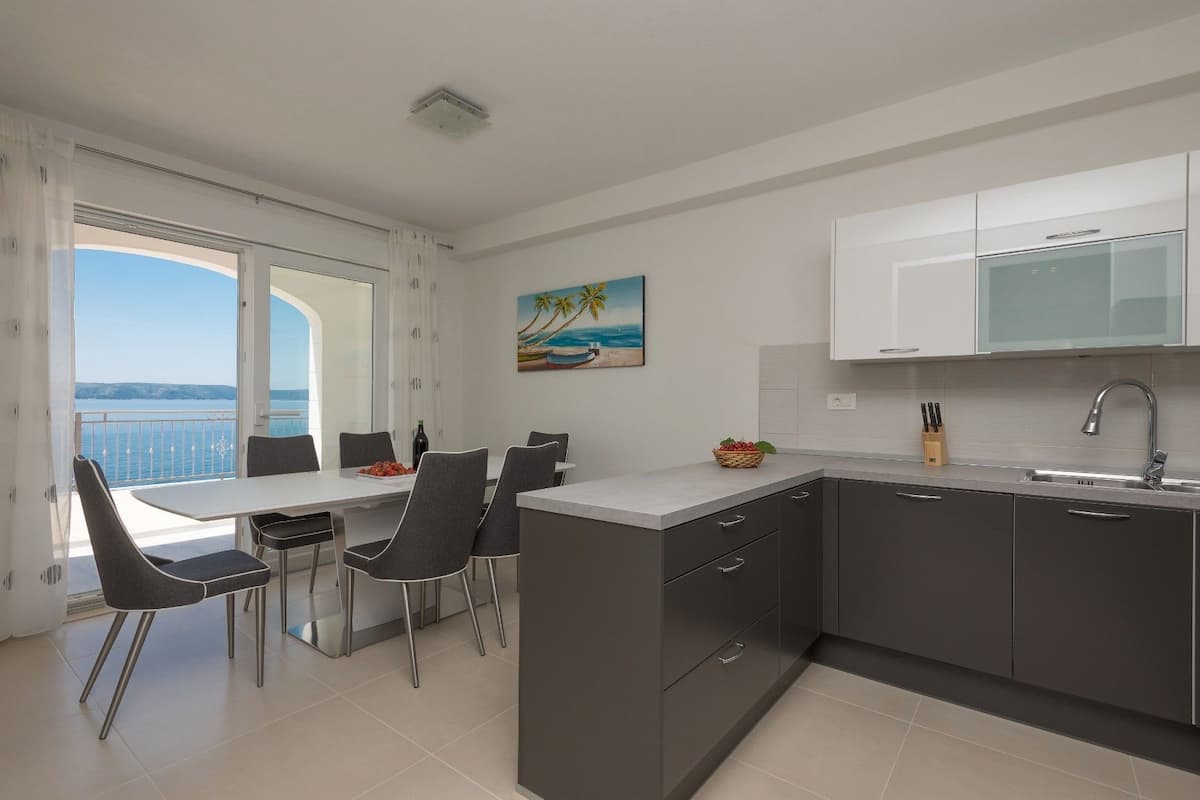 A modern kitchen with dark cabinets, a white countertop, and a view of the sea through a large window.