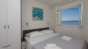 Un lit bien fait, avec des draps blancs, une couverture grise et une serviette pliée posée sur le sol. Au mur, au-dessus du lit, on peut voir un tableau représentant une fleur bleue.