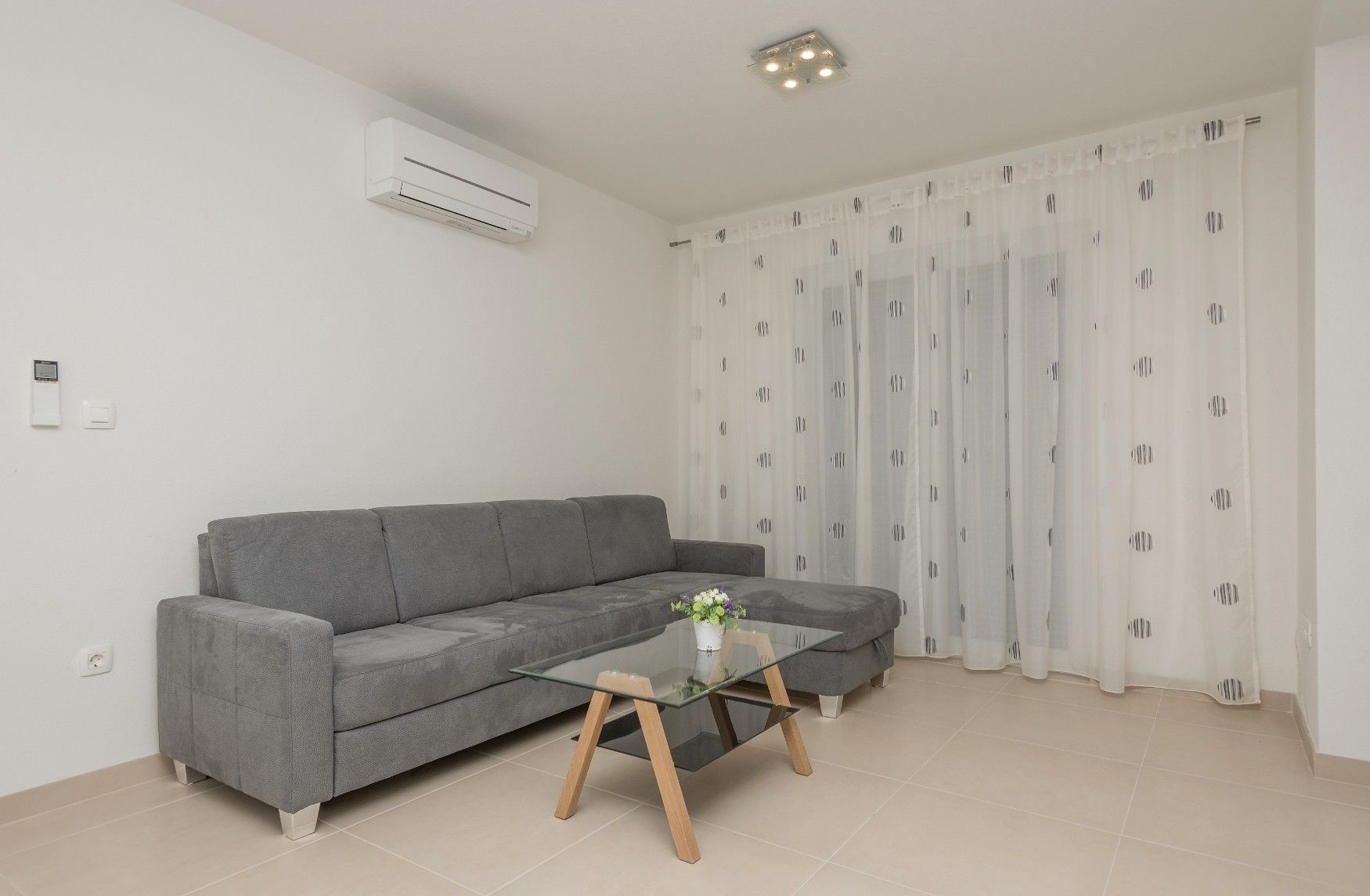 Een moderne woonkamer met een grijze bank, een glazen salontafel en een witte airconditioner aan de muur.