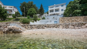 Un hôtel situé sur la côte, avec un mur en pierre, une eau cristalline et des bâtiments modernes en arrière-plan.