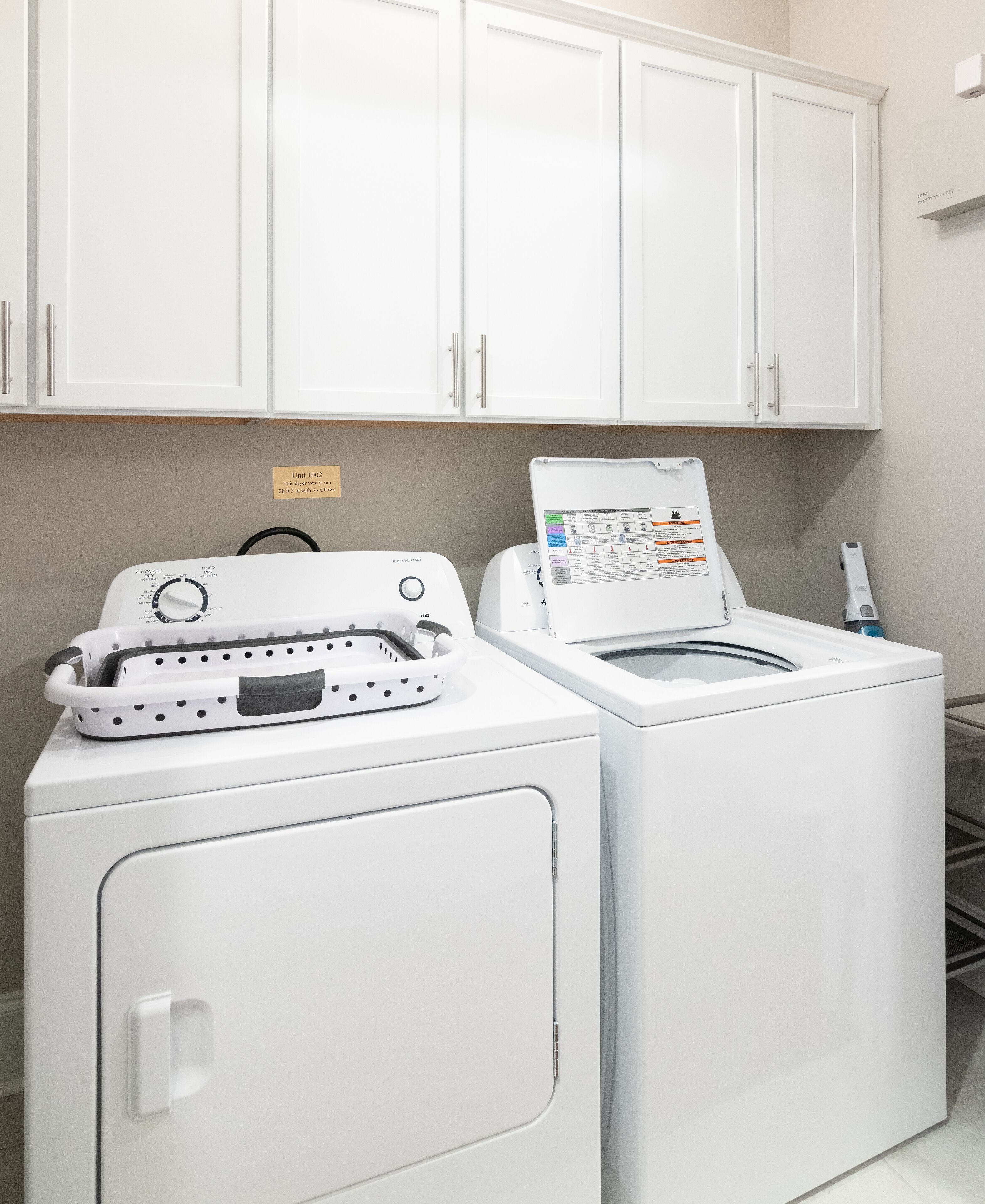 Een wasruimte met twee witte wasmachines, waarvan er een bedieningspaneel heeft en de andere een zeepdispenser. Daarboven hangt een bord met het appartementsnummer.