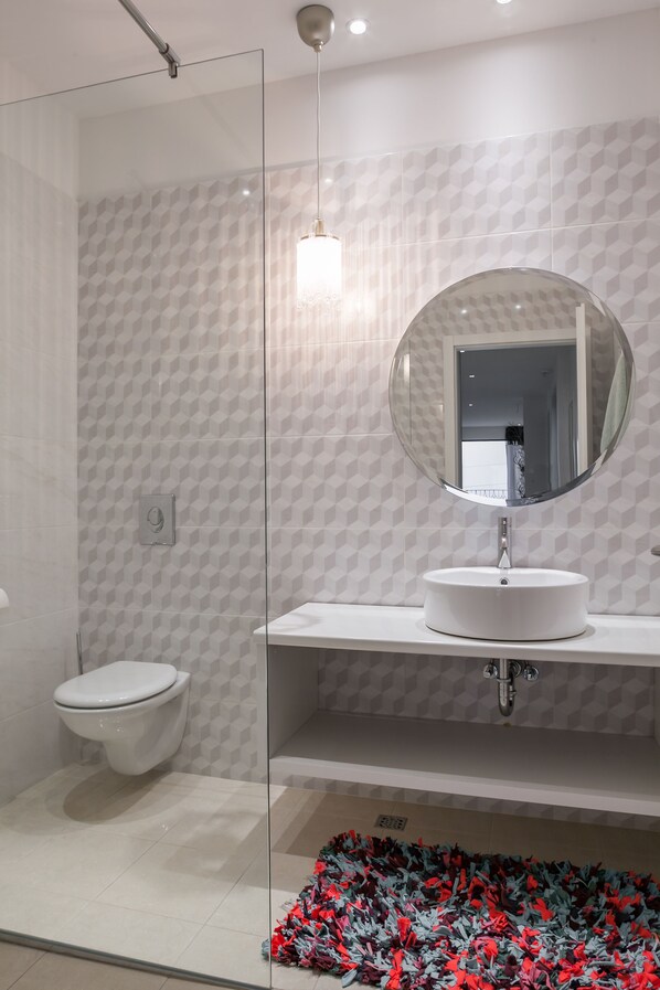 A modern bathroom with a glass shower enclosure, a toilet, a sink with a round mirror, and patterned wall tiles. - Villa Tres Chic (Makarska)
