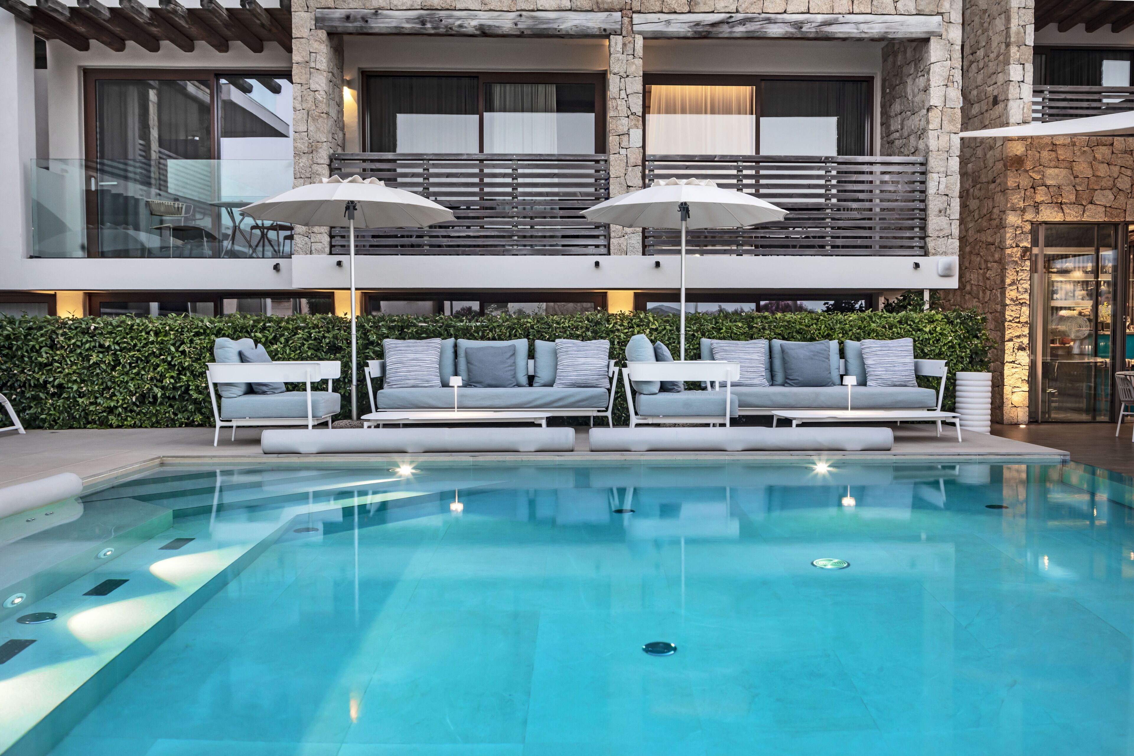 A modern pool area with two lounge chairs and umbrellas, a stone building with balconies, and a clear blue pool.
