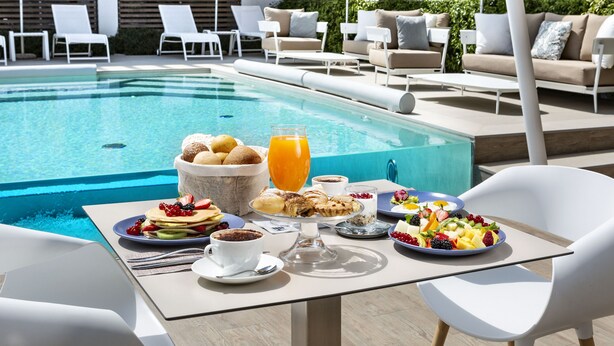Un coin petit-déjeuner aménagé au bord de la piscine, avec une table, des chaises et une variété de plats et de boissons.