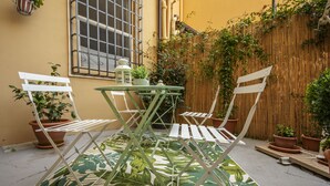 Un patio con una mesa redonda y sillas blancas, rodeado de plantas en macetas y una valla de bambú.