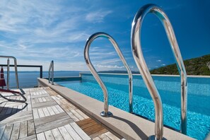 A poolside area with clear blue water, metal poolside railings, and a wooden deck. - Villa Vilovic (Makarska)