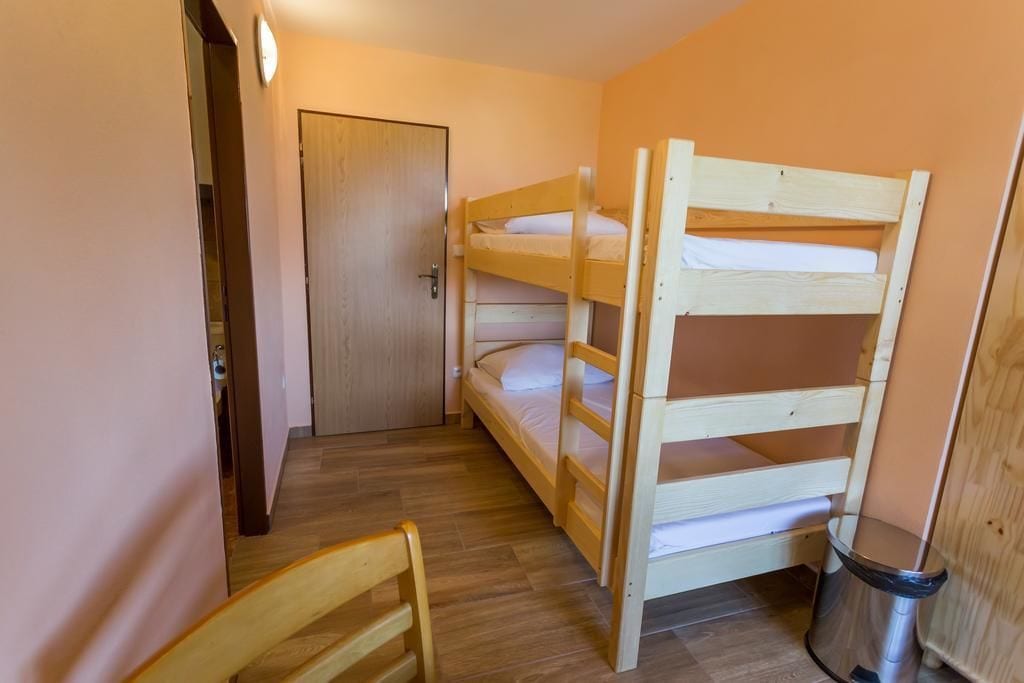 Habitación con literas, una puerta de madera, un cubo de basura y una silla de madera.