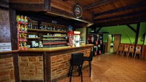 Zona de bar con mostrador de ladrillo, estanterías con botellas, vasos y aperitivos. Hay sillas y una pared verde al fondo.