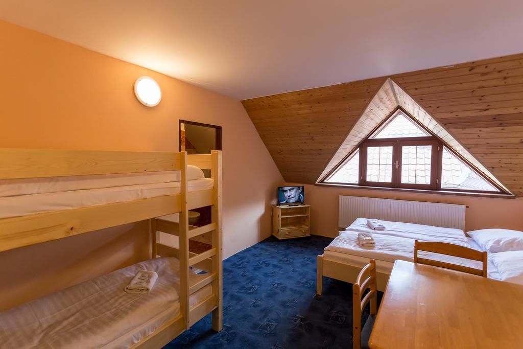 A dormitory room with bunk beds, a small table, a chair, a window with a triangular wooden ceiling, and a wall-mounted light.