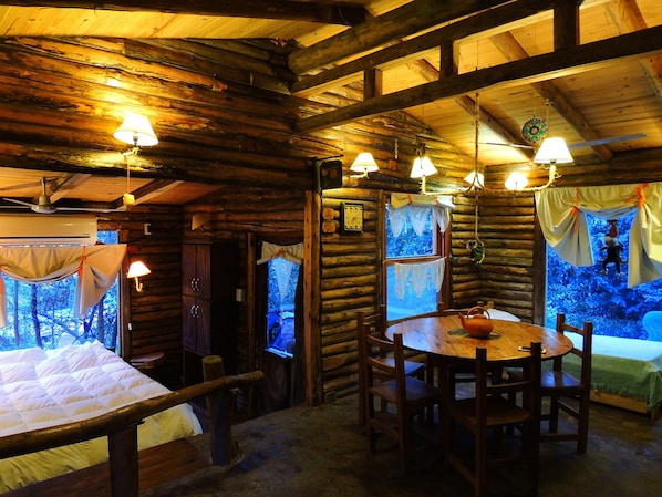 Interior acogedor de una cabaña de troncos con una mesa redonda de madera, sillas y una cama con colcha blanca.