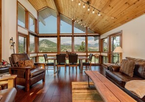 A spacious room with a high ceiling, large windows offering a mountain view, wooden furniture, and a dining area.