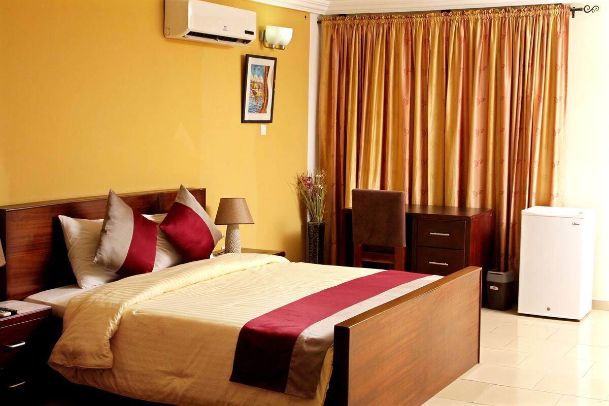 A hotel room with a bed, a desk, a chair, a lamp, a trash bin, and a refrigerator.
