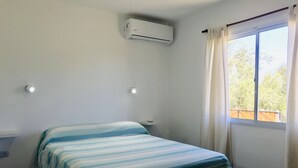 Una habitació amb un llit, un aparell d'aire condicionat, una tauleta de nit i una finestra amb cortines.