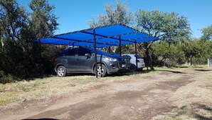 Un cotxe aparcat sota una marquesina blava en un entorn natural amb arbres i un camí de terra.