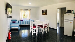 Habitació amb dos llits, una taula de menjador amb cadires, un televisor fixat a la paret, un refrigerador i un extintor.