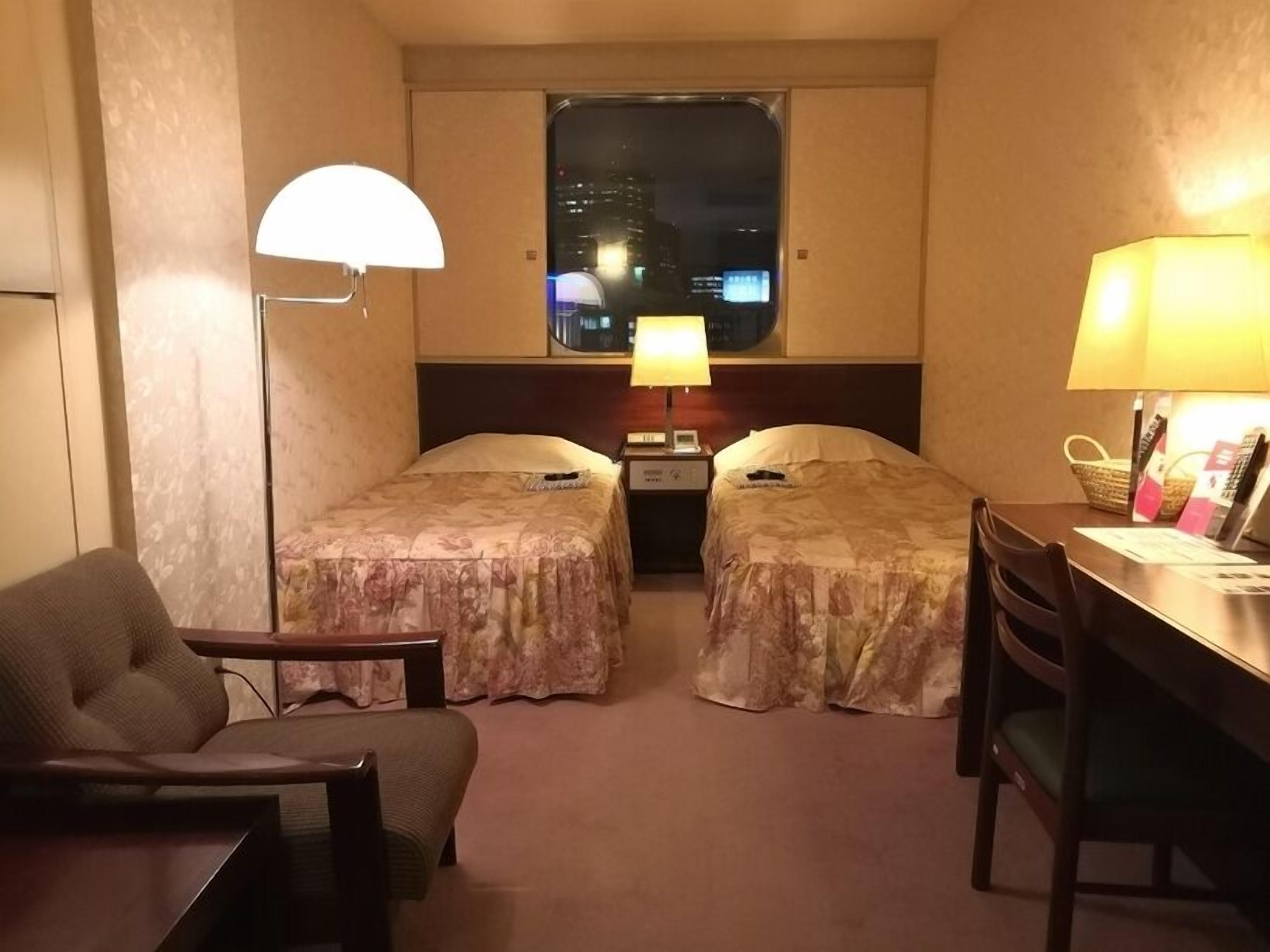 A hotel room with two beds, a desk, a chair, a lamp, and a window with a city view.