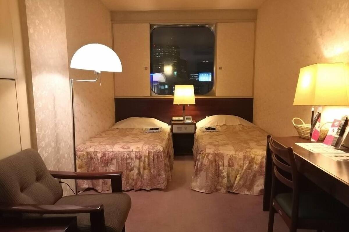 A hotel room with two beds, a desk, a chair, a lamp, and a window with a city view.