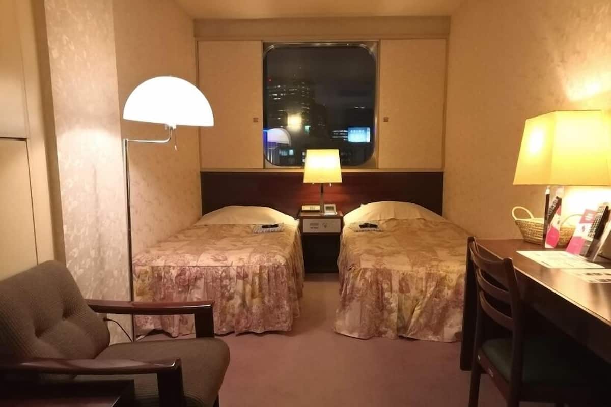 Ein Hotelzimmer mit zwei Betten, einem Schreibtisch, einem Stuhl, einer Lampe und einem Fenster mit Blick auf die Stadt.