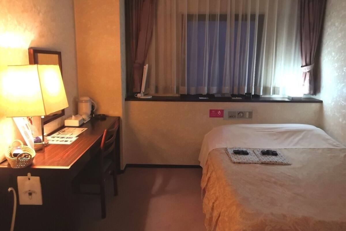 A hotel room with a desk, a lamp, a bed, a window with curtains, and a wall-mounted control panel.