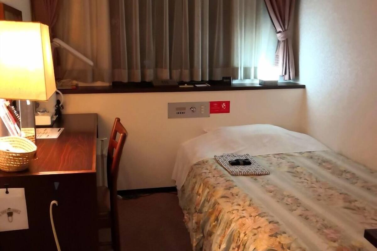 A hotel room with a bed, a desk with a lamp, a chair, a window with curtains, and a control panel.