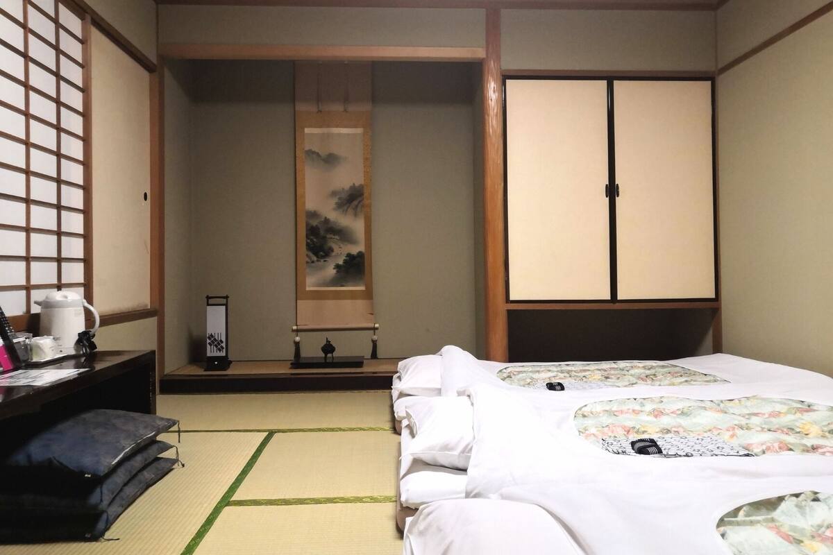 A traditional Japanese-style room with tatami flooring, a bed with a floral bedspread, a desk with a kettle, and a sliding door closet.