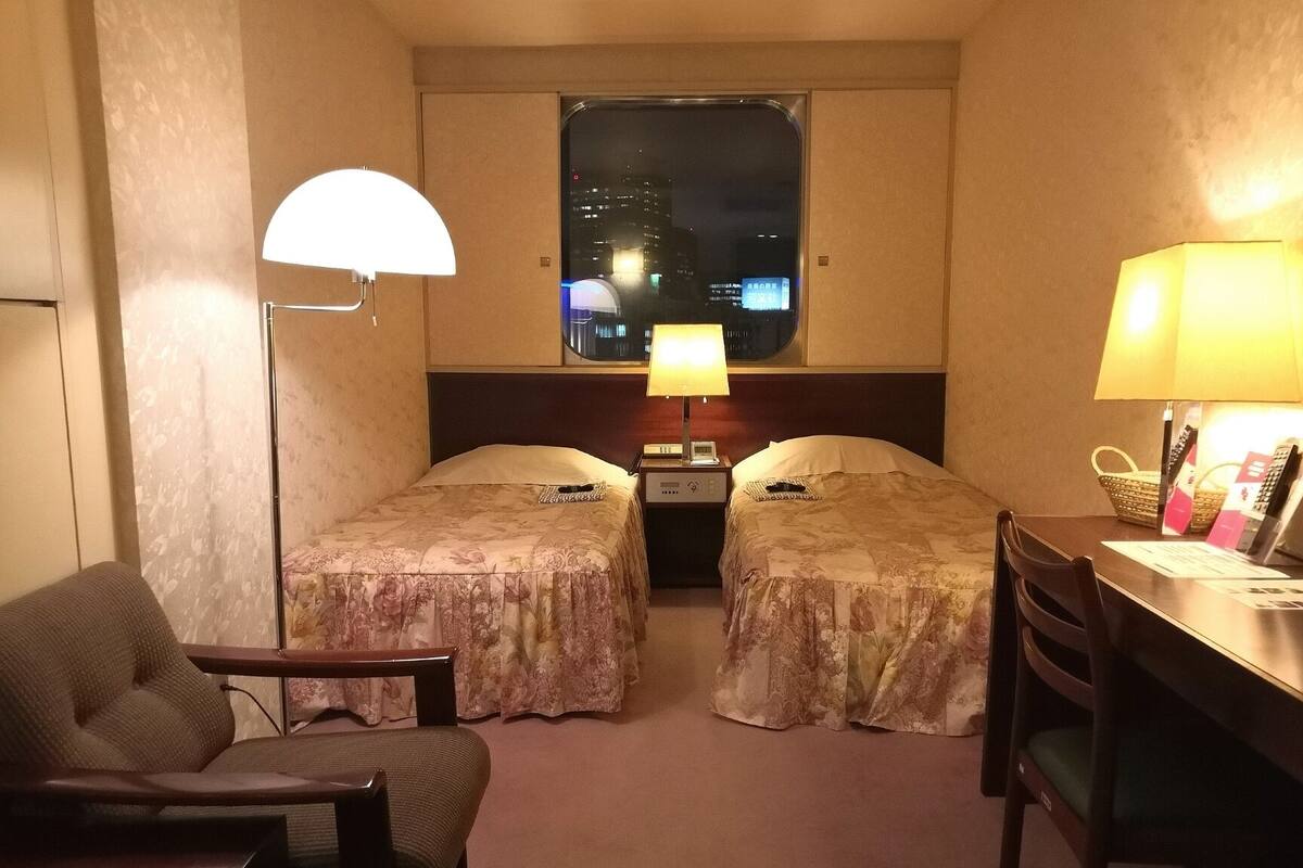 A hotel room with two beds, a desk, a chair, a lamp, and a window with a city view.