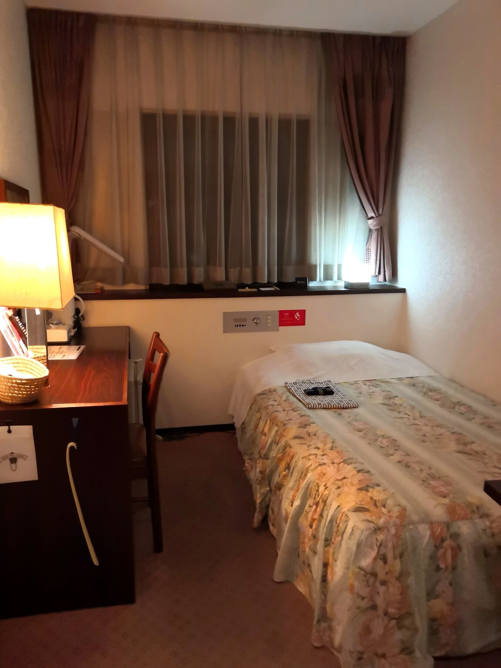 Ett hotellrum med en säng, ett skrivbord med en lampa, en stol, ett fönster med gardiner och en kontrollpanel.