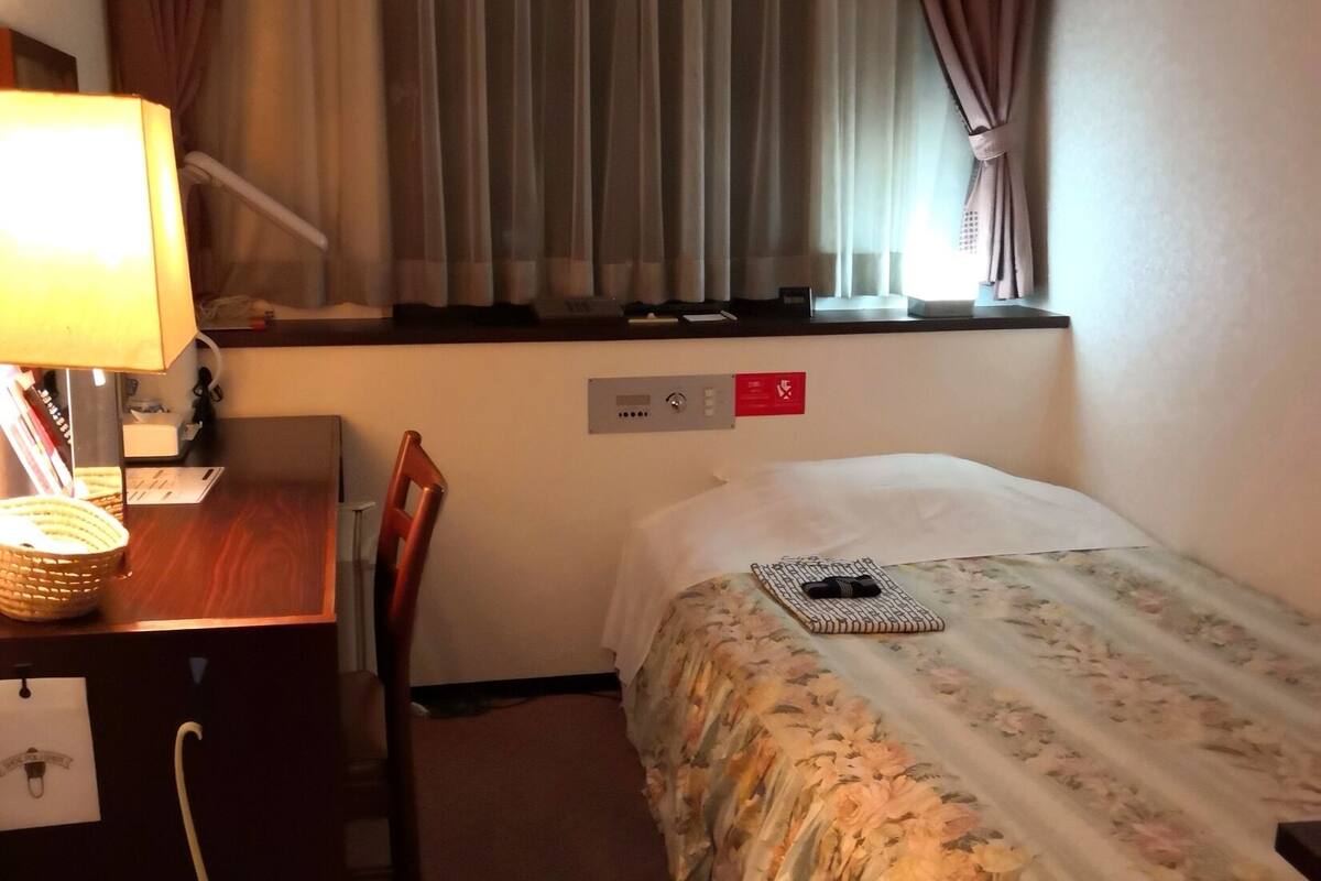 A hotel room with a bed, a desk with a lamp, a chair, a window with curtains, and a control panel.