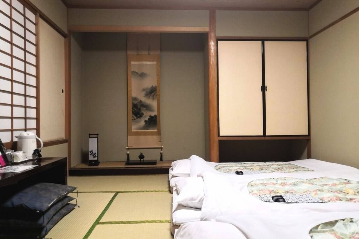 A traditional Japanese room with tatami flooring, a bed with a floral bedspread, a desk with a kettle, and sliding doors.