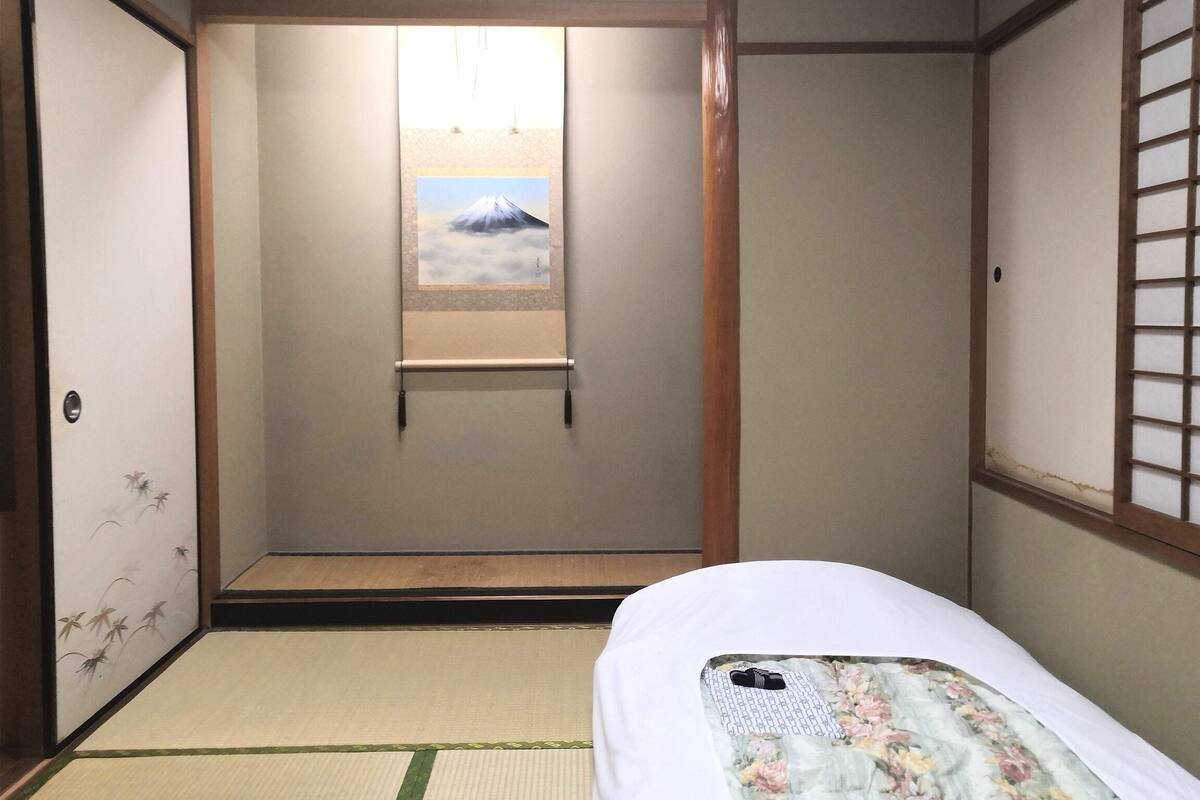 A traditional Japanese room with tatami flooring, a shoji screen, a bed with a floral bedspread, and a framed picture of a mountain.