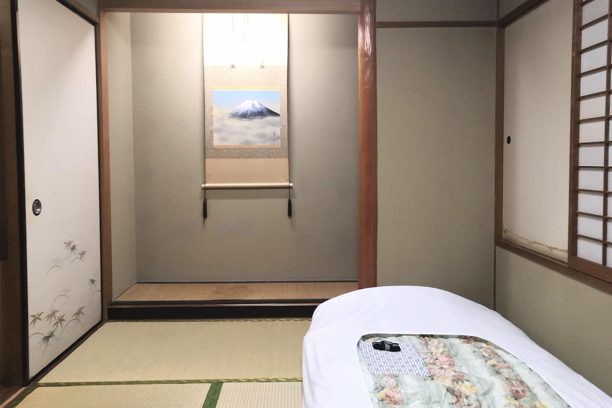 Ein traditioneller japanischer Raum mit Tatami-Boden, Shoji-Schirm und einem Bett mit floralem Überwurf. Es ist ein gerahmtes Bild einer Berglandschaft zu sehen.
