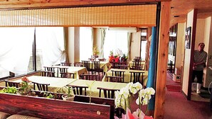 Un restaurante con mesas y sillas de madera, plantas en macetas y una persona de pie en el umbral.