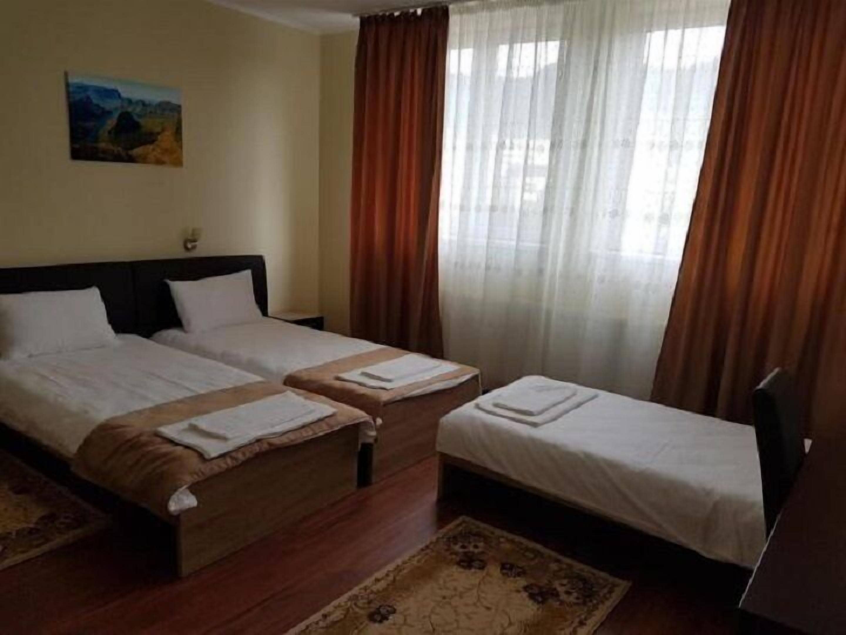 A hotel room with two single beds, a wooden floor, a rug, a painting on the wall, and a window with curtains.