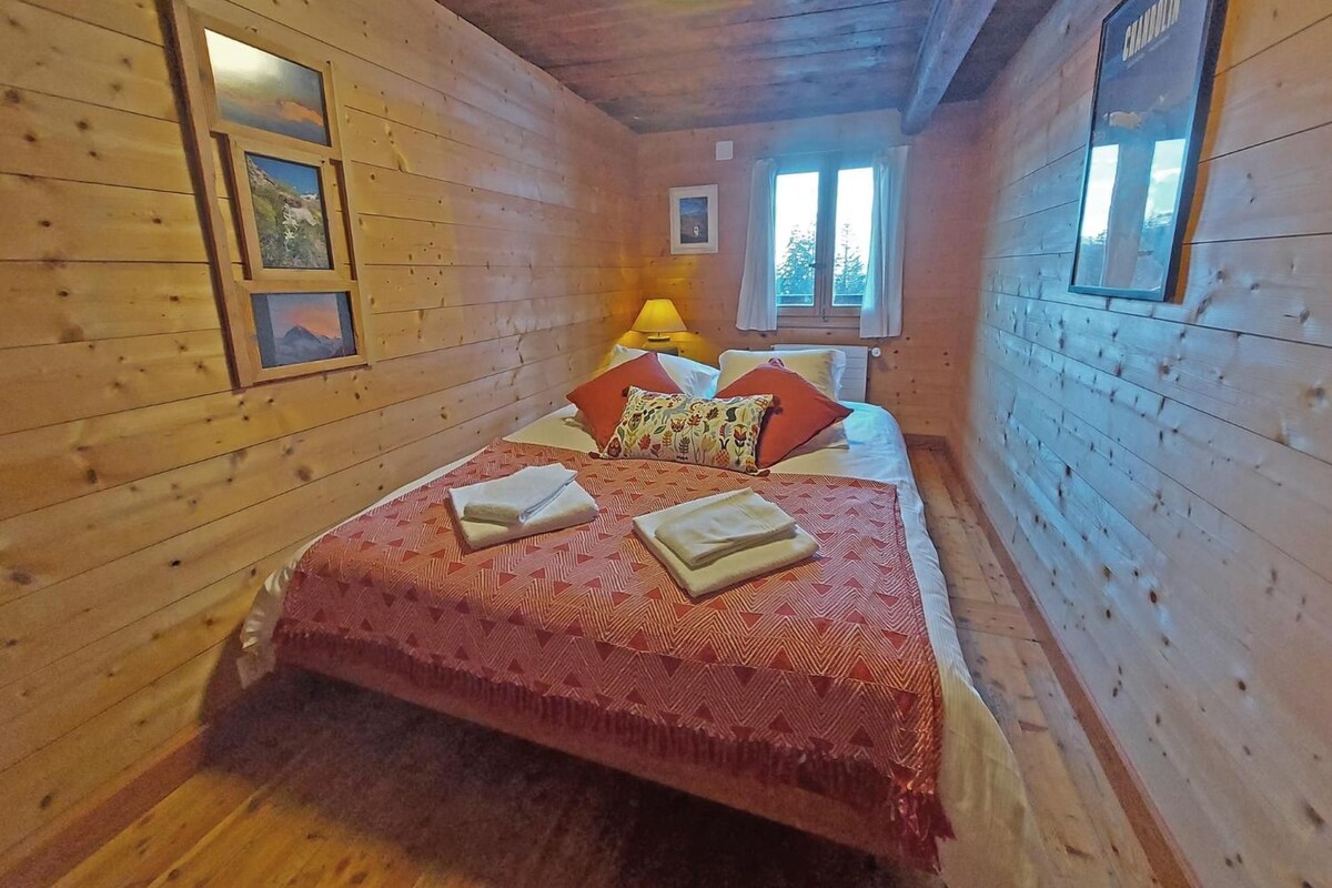 Habitación de cabaña de madera con una cama, dos ventanas, una lámpara y cuadros en las paredes.