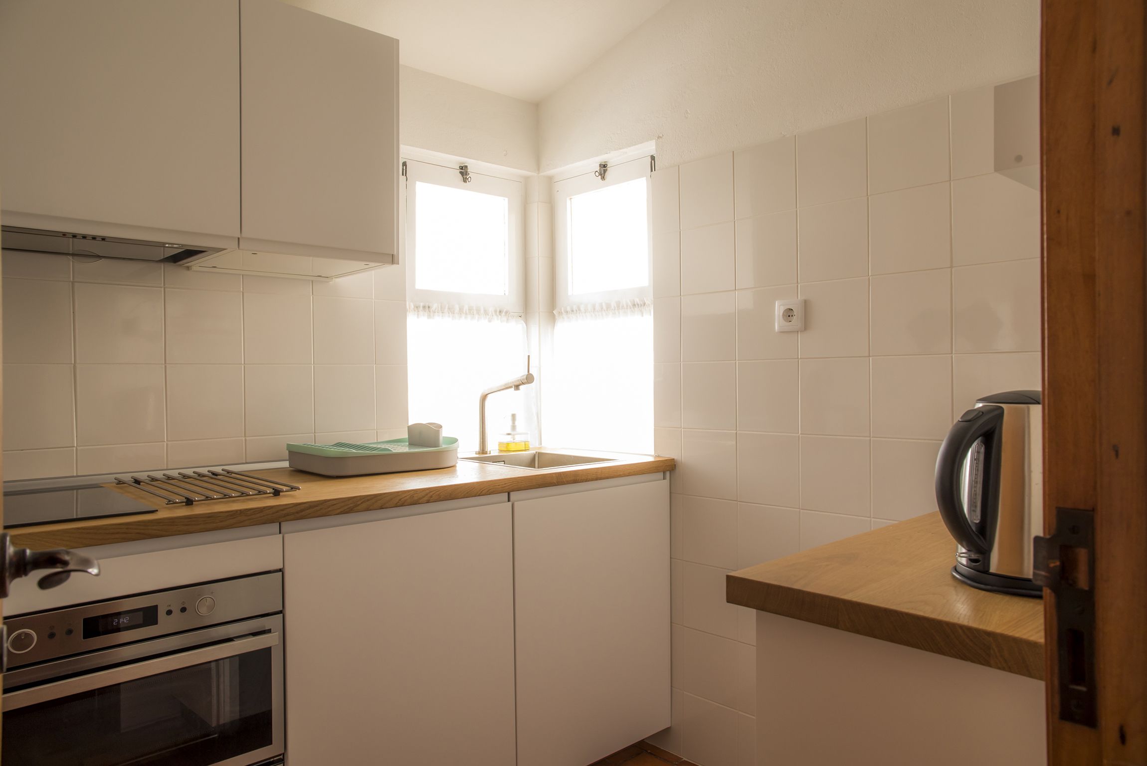 Una cocina compacta con armarios blancos, horno integrado, lavamanos con ventana encima y hervidor en la encimera.