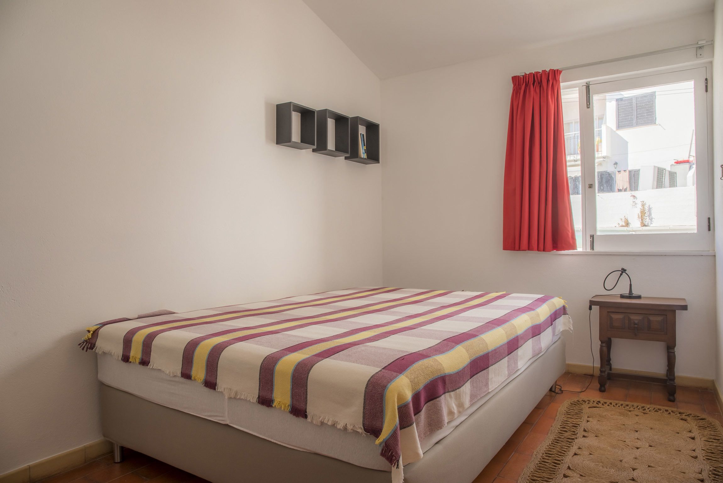 A bedroom with a bed, a bedside table, a lamp, a window with curtains, and a wall-mounted shelf with books.
