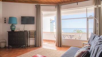A room with a wooden ceiling, a TV, a sofa, a red cushion, a lamp, a dresser, and a view of the sea through large windows.