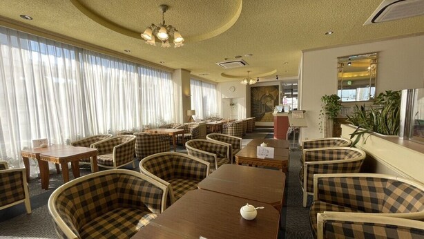 Un espace de restauration d’hôtel avec des chaises à motifs à carreaux et des tables en bois, doté d’un plafond avec des luminaires encastrés et d’une grande fenêtre agrémentée de rideaux.
