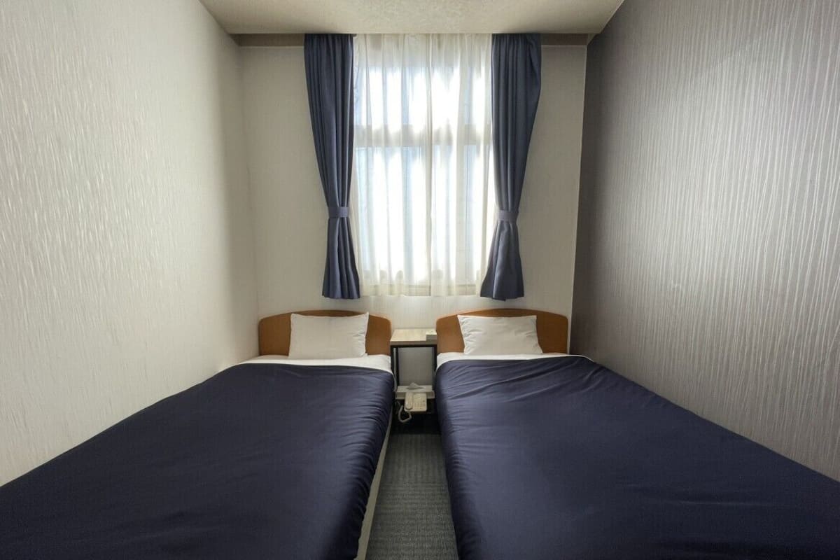 A hotel room with two single beds, a small bedside table, and a window with curtains.