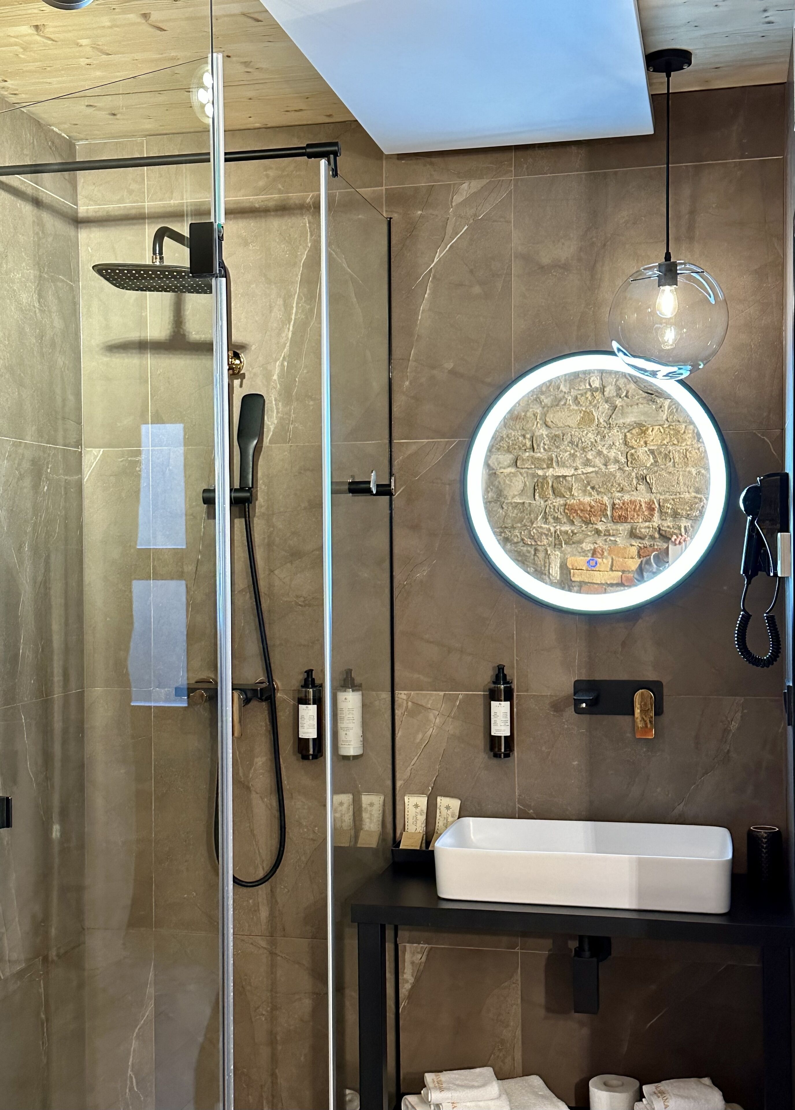 Un baño moderno con ducha de vidrio, un espejo redondo y un diseño de pared de ladrillo, y un lavabo blanco con encimera negra.