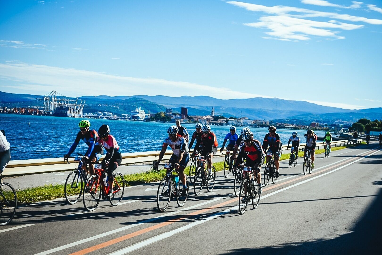Un grupo de ciclistas en una carretera, con una masa de agua y estructuras industriales al fondo.