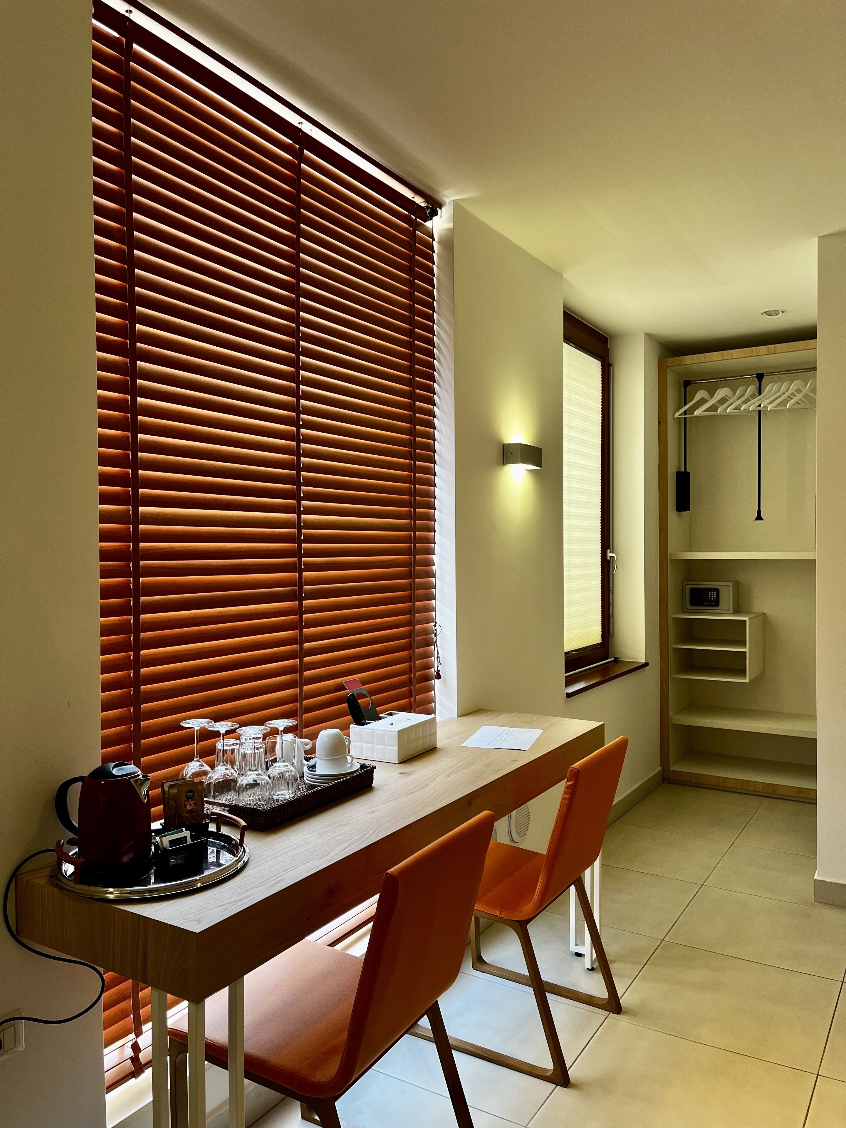 A dining area with a wooden table, two orange chairs, a tray with glasses and a kettle, and a wall-mounted light.