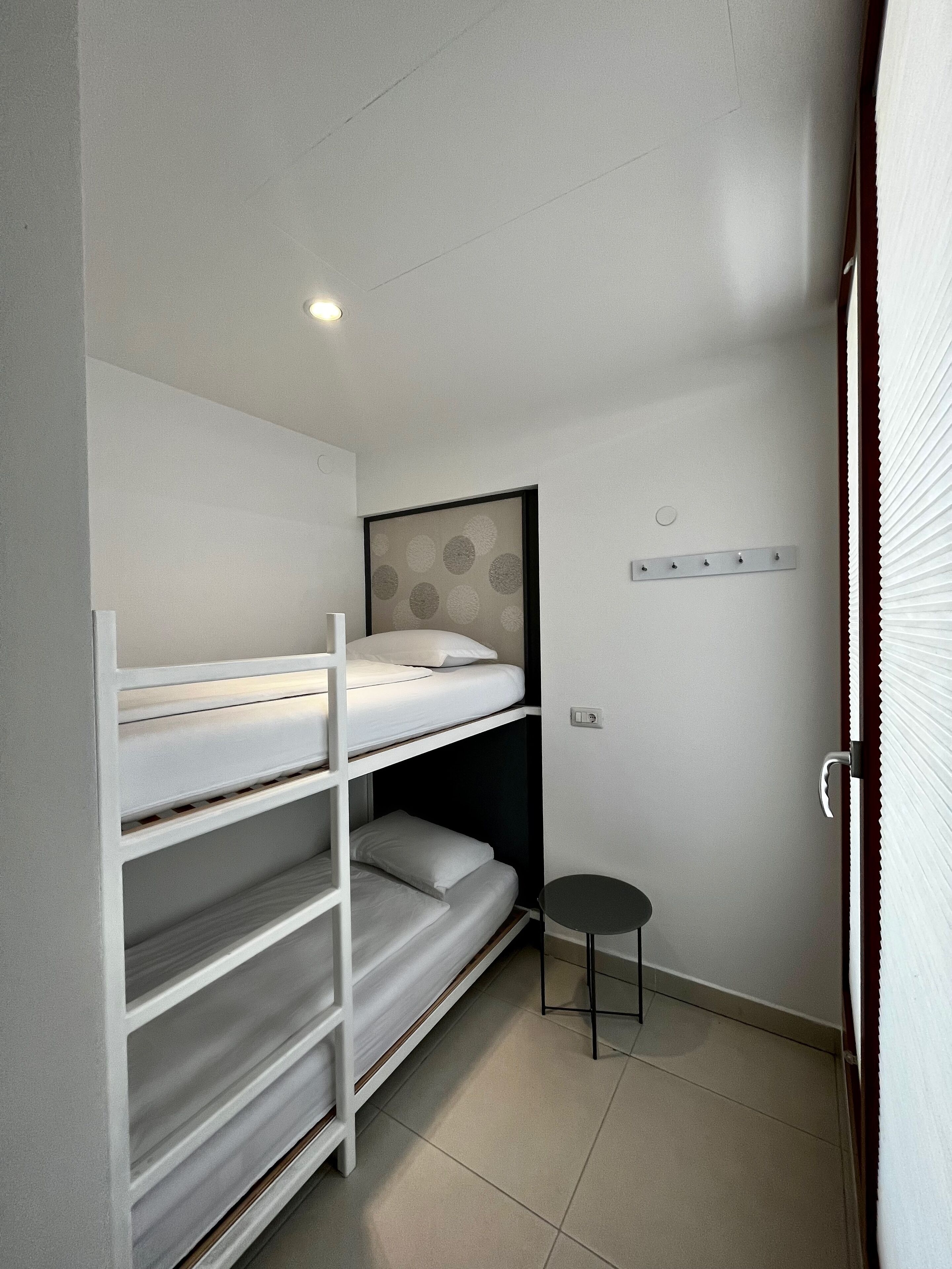 Quarto compacto com beliches, uma mesinha e luminária na parede.
