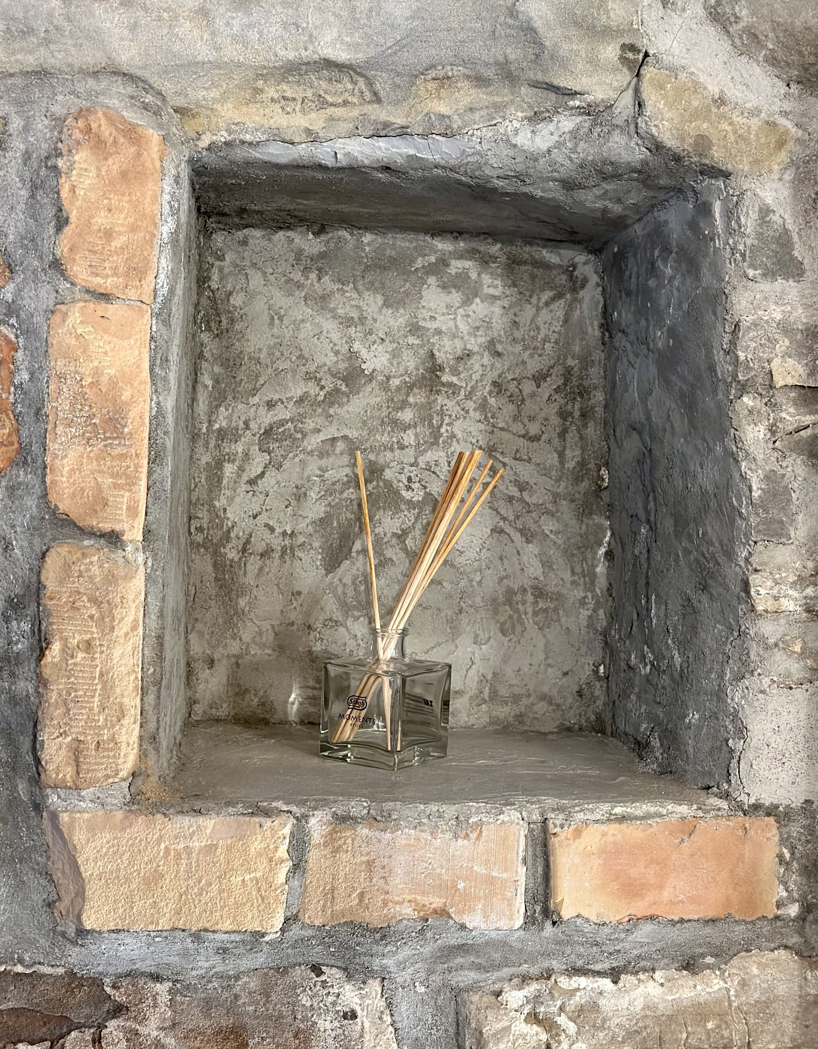 Un camino in pietra con un'apertura quadrata, un vaso di vetro con bastoncini di canna e un piccolo ornamento appeso.