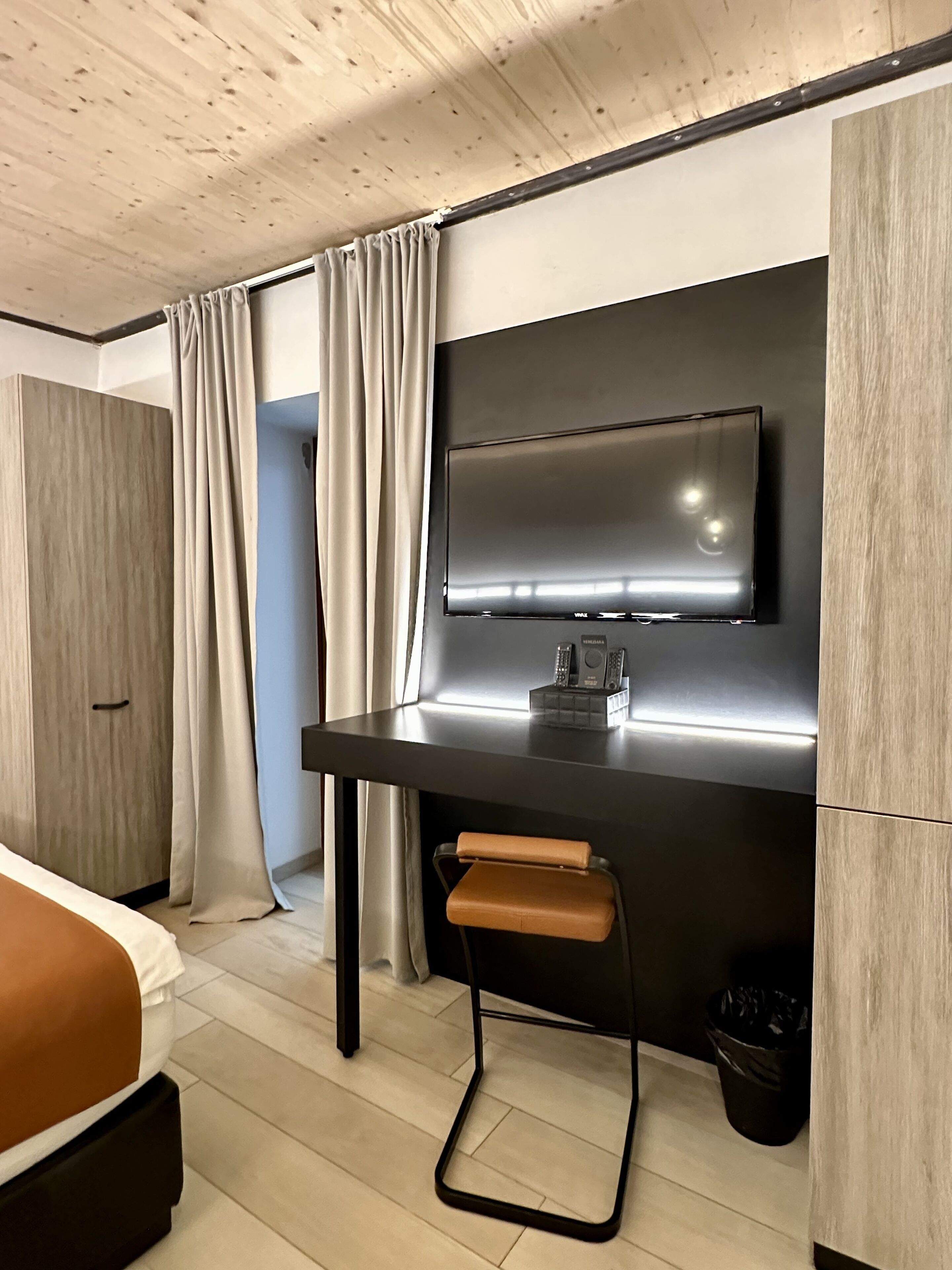 A modern hotel room with a wooden ceiling, a large bed with a brown headboard, a desk with a flat-screen TV, a small stool, and a wardrobe.