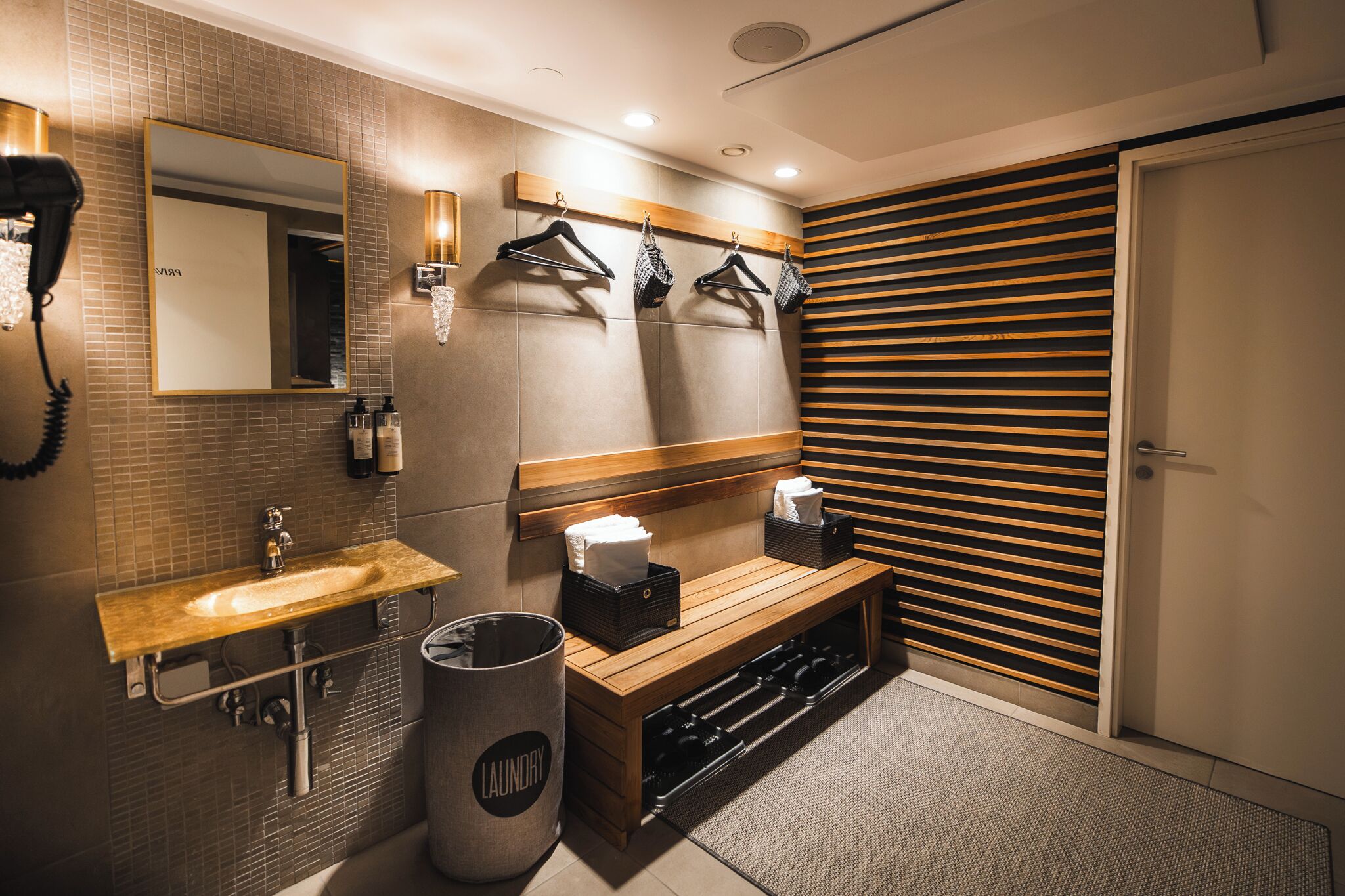 A modern bathroom with a sink, mirror, towel rack, bench, and a wall-mounted towel dispenser.