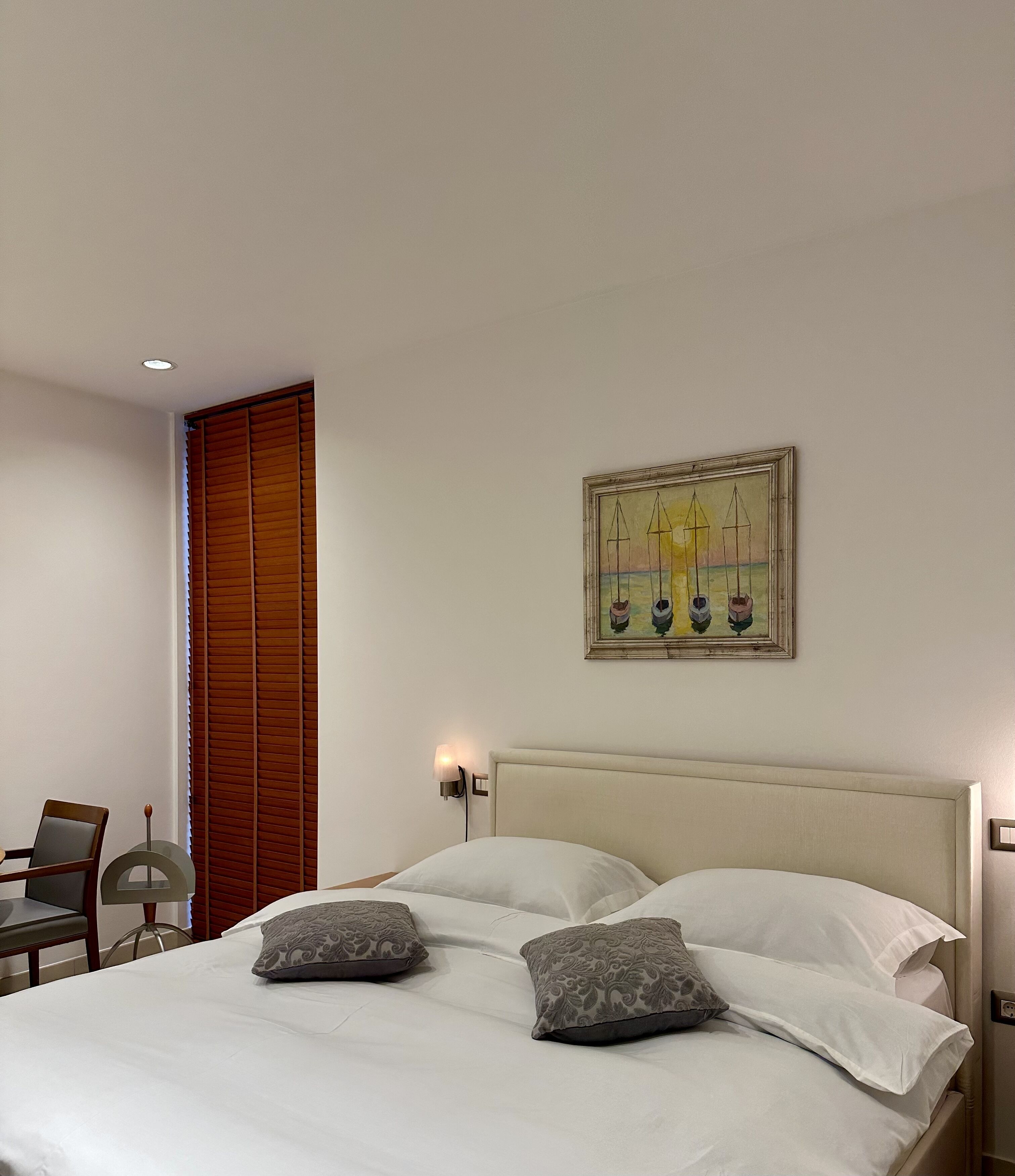 Una cama bien hecha con dos almohadas, una silla de madera, un cuadro colgado en la pared y una mesita de noche con una lámpara.