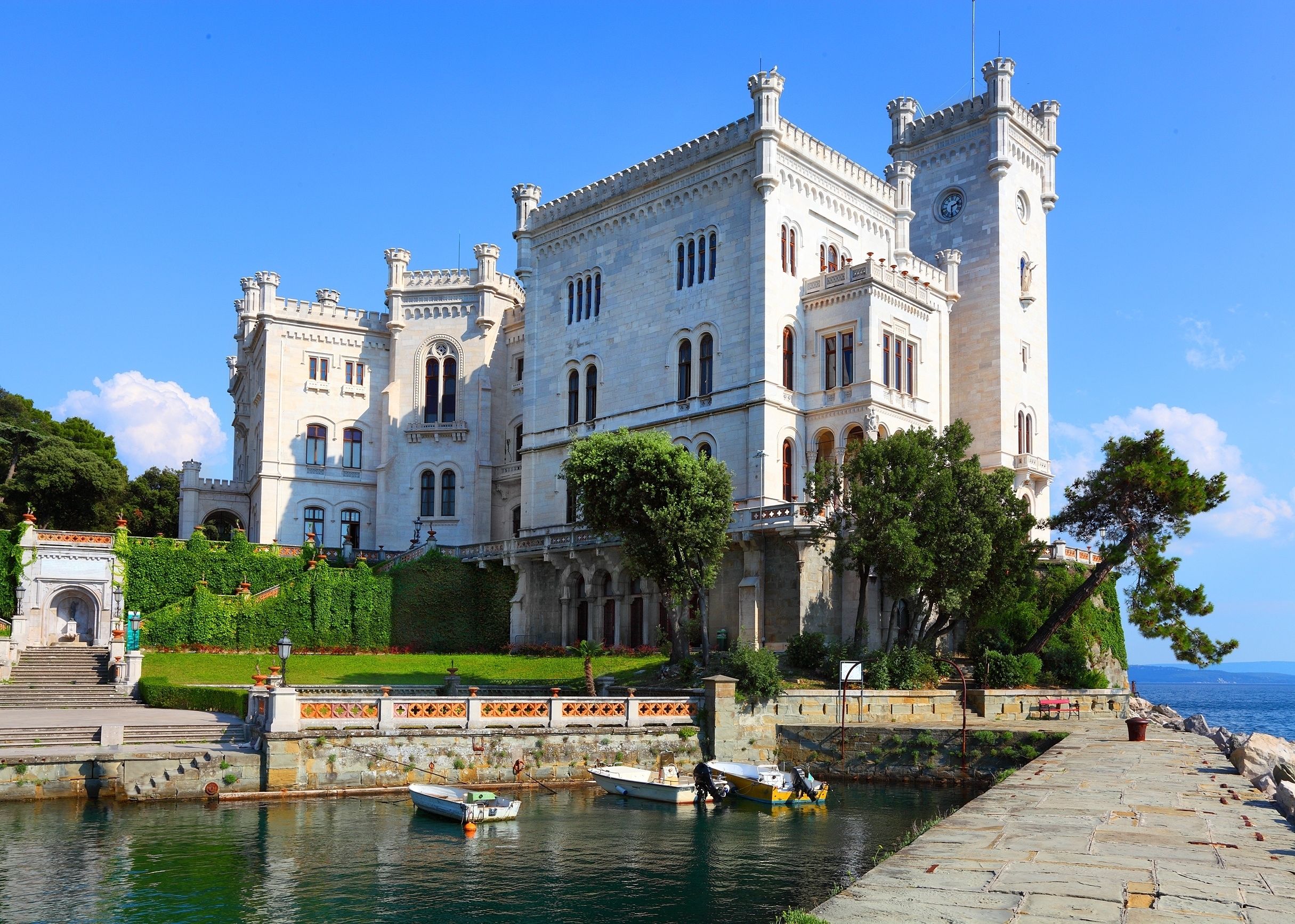A large, historic building with a prominent tower and arched windows, situated by a waterfront with boats docked.