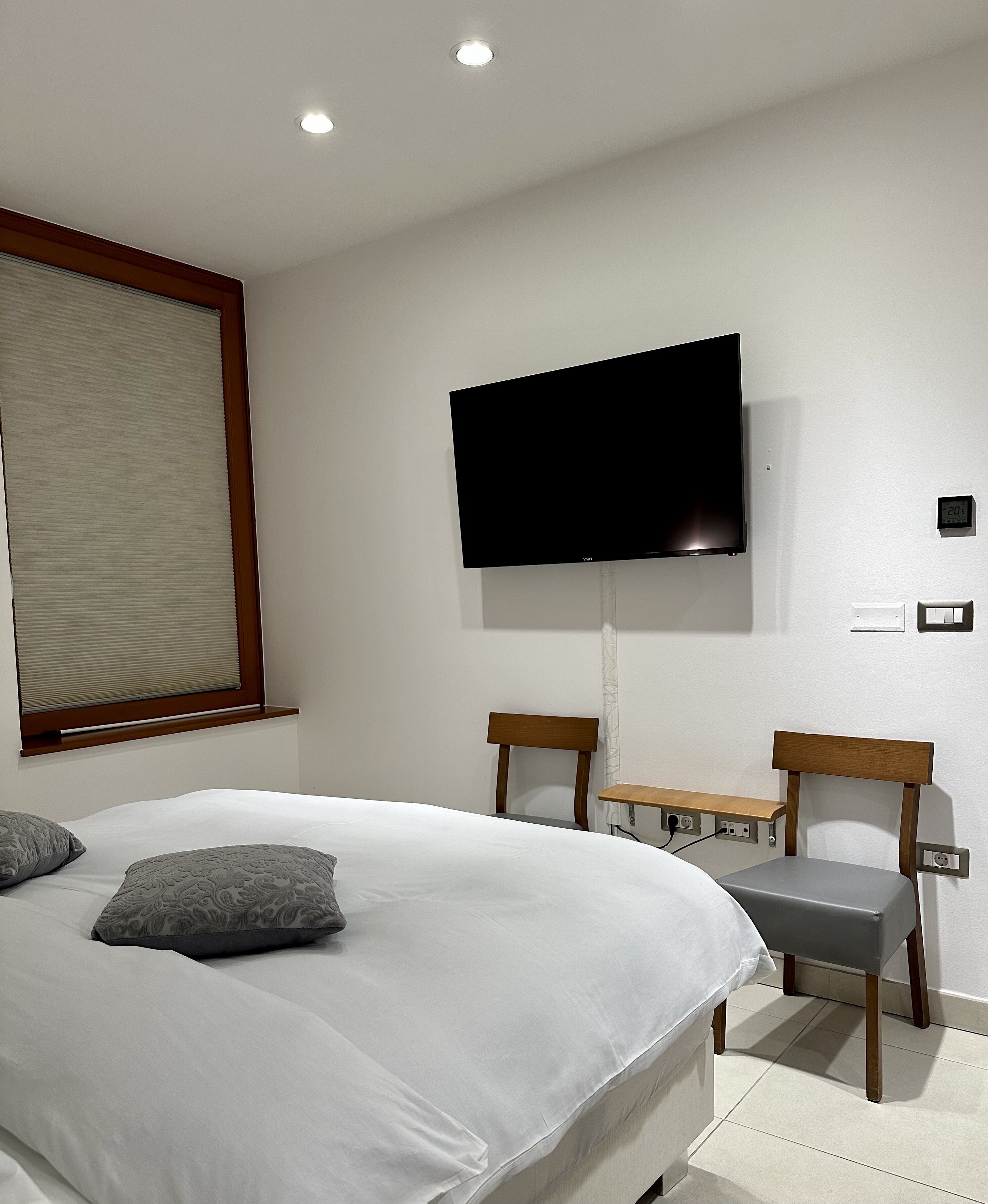 Un dormitorio con una cama, televisión montada en la pared, una mesita, dos sillas y una ventana con persianas.