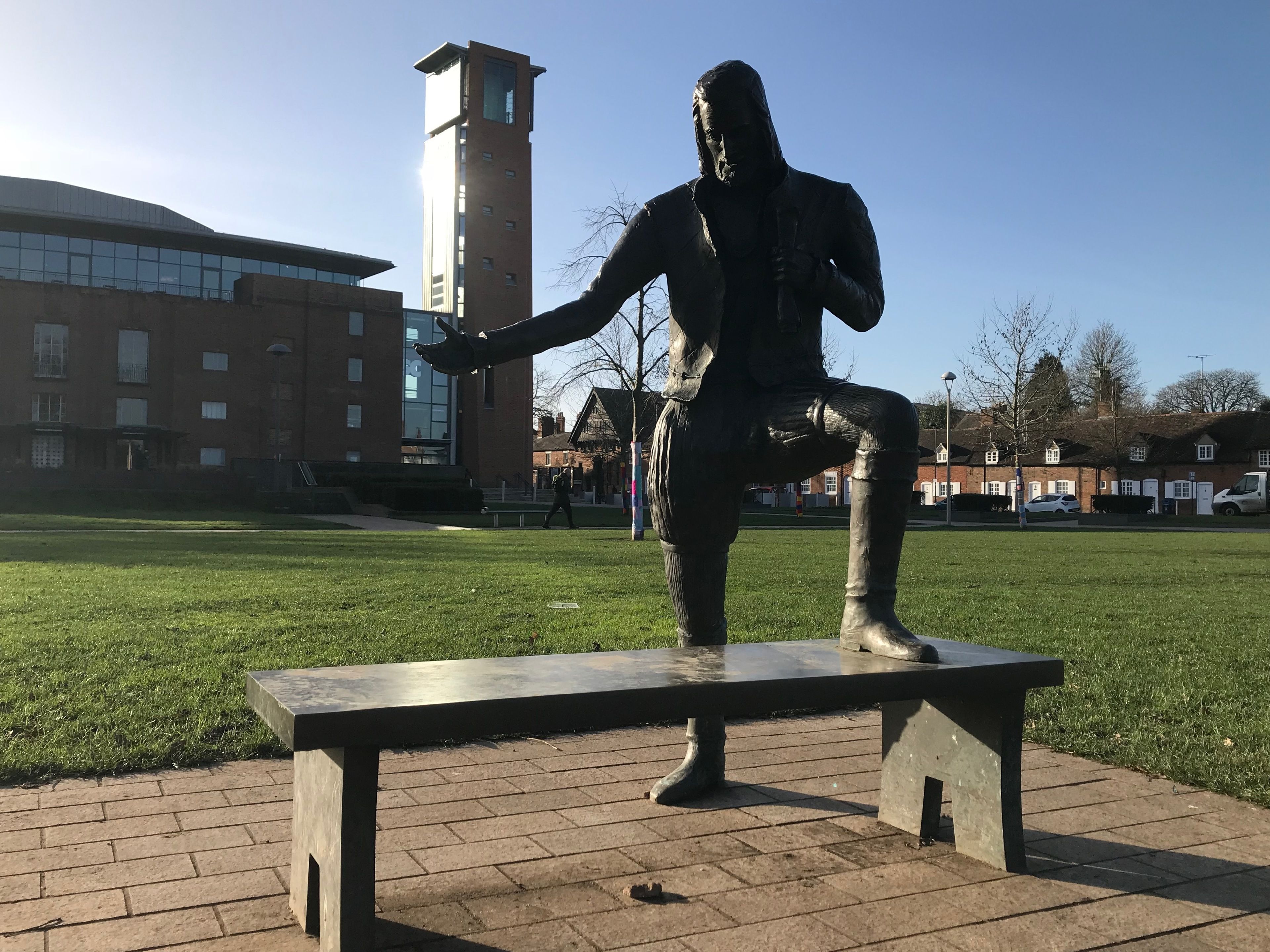 A statue of a person seated on a bench in a park with a building in the background.